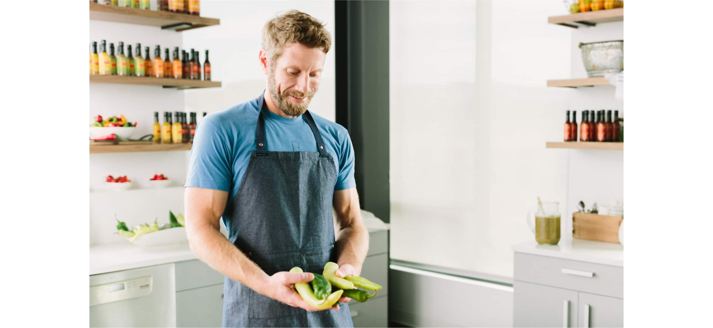 John standing in his kitchen holding peppers in his hands, with rows of small sauce bottles lining the shelves behind him.